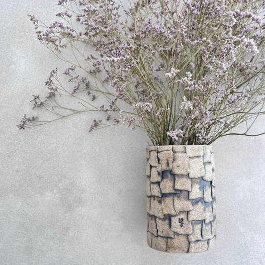 A handcrafted ceramic vase with a geometric pattern, predominantly beige in color, containing a bundle of small purple flowers.